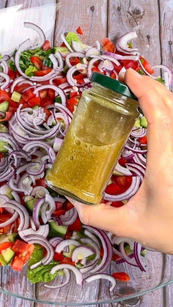 Dressing für low carb Brotsalat in einem Schraubglas über einem Brotsalat mit Tomaten, roten Zwiebeln, Paprika und Gurken in einer Glasschale