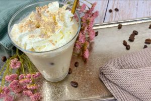 Protein Eiskaffee mit Sahne, Eiswürfeln und Chunky Flavour im Glas auf einem Tablett mit Blumen und Kaffeebohnen