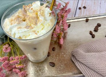 Protein Eiskaffee mit Sahne, Eiswürfeln und Chunky Flavour im Glas auf einem Tablett mit Blumen und Kaffeebohnen